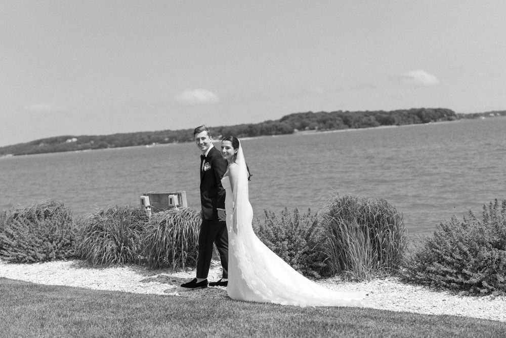 Black and white wedding photo by the water with couple in classic attire.