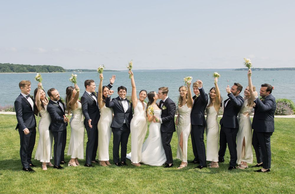 Wedding party in navy and white poses cheerfully on oceanfront lawn.