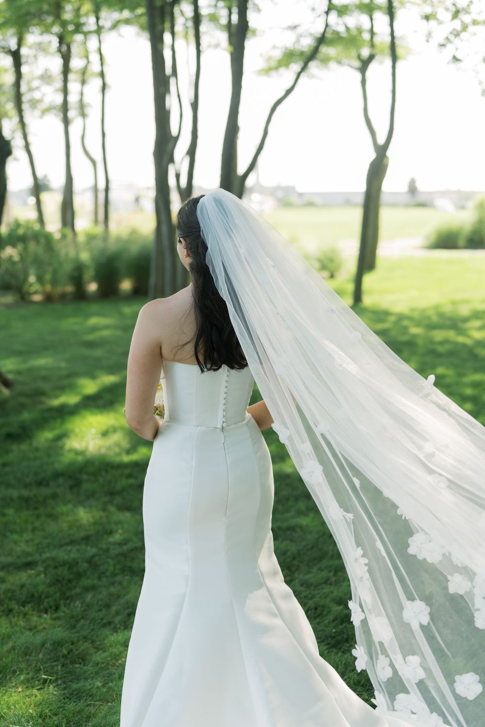 A flowing white wedding veil catches the breeze in a sunlit garden setting.