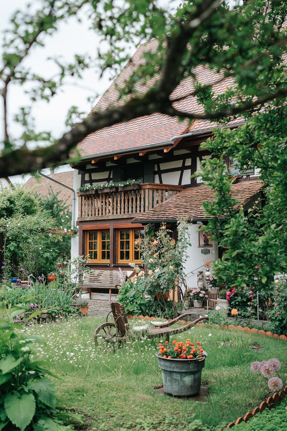Charming European cottage house with timber frame details, balcony, and colorful garden featuring potted flowers and rustic decor.