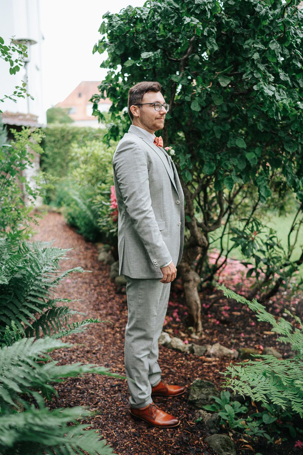A person stands on a mulch path in a lush garden wearing a light gray suit with brown shoes and a bow tie.