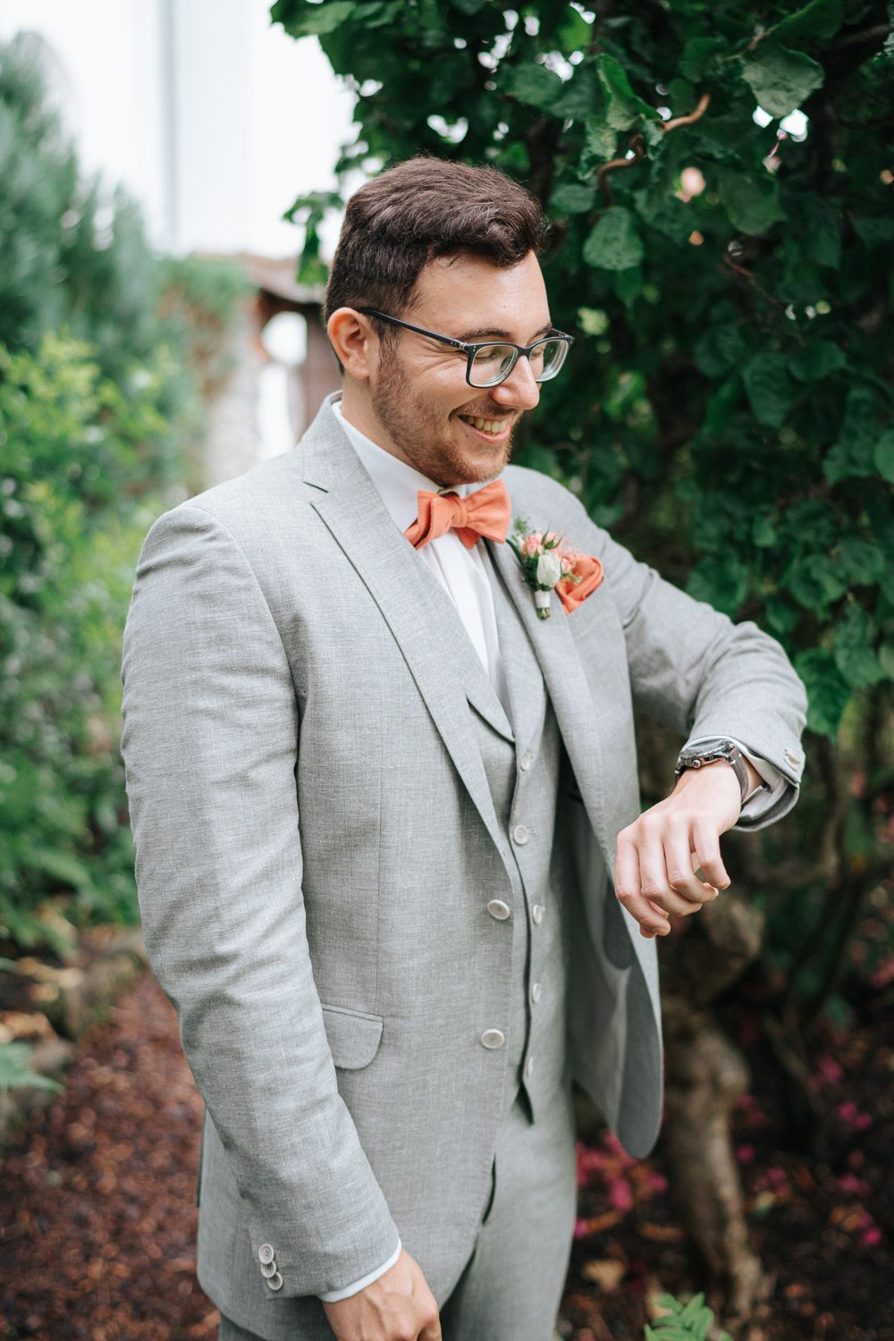 A person in a gray suit with coral-colored bowtie looks at their watch in a garden setting.