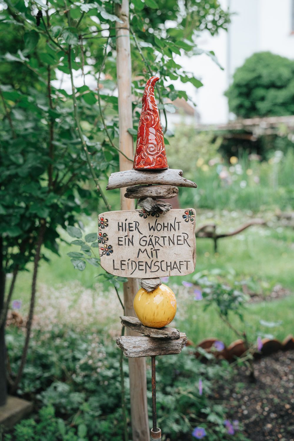A decorative garden pole displays a German sign with a Coca-Cola bottle cap on top amidst green foliage.
