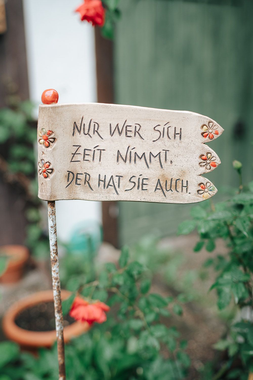Three wooden garden markers display a German phrase across them among green foliage and red flowers.