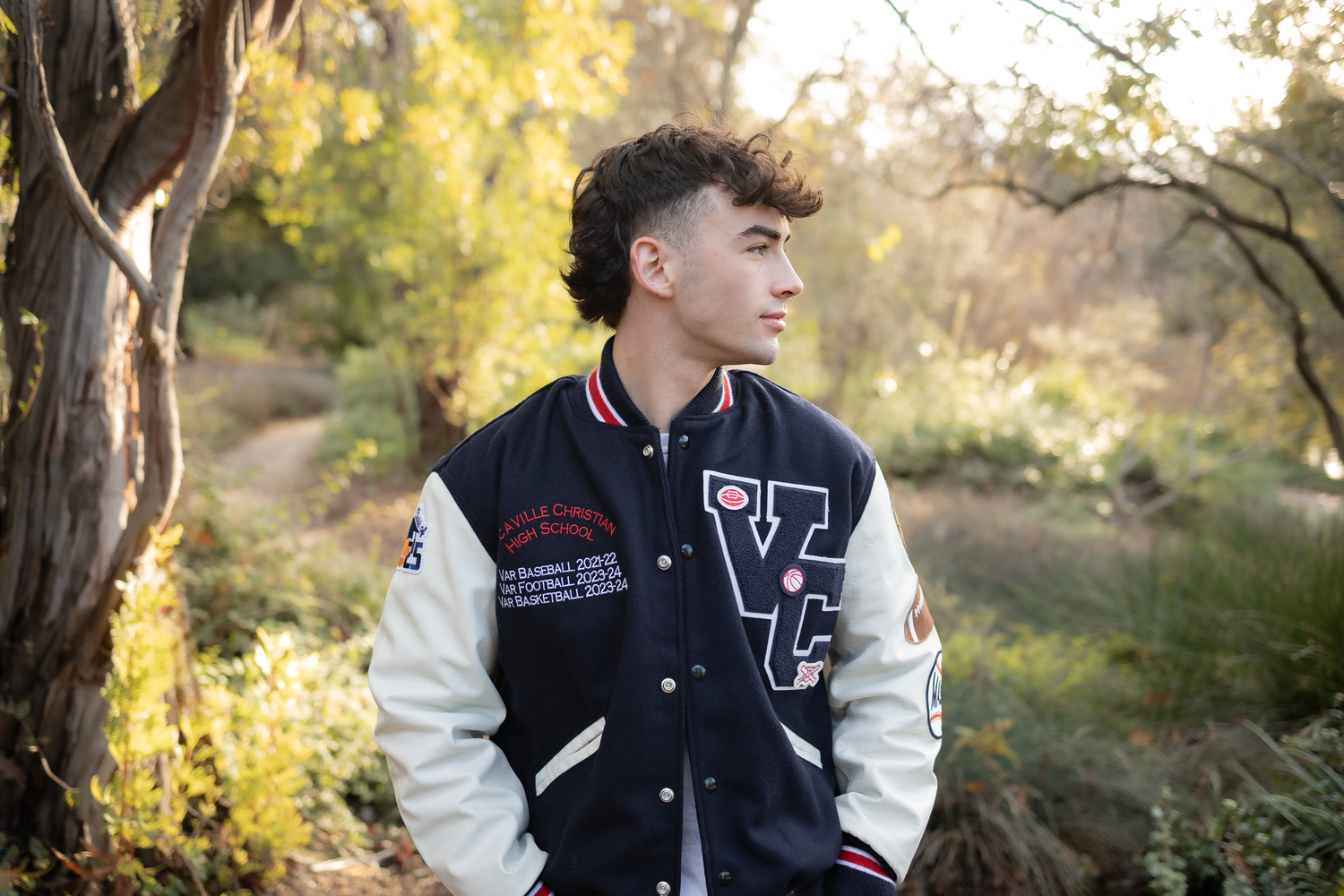 Senior portrait of white male student in letterman jacket in a garden, staring to the side with a stoic expression