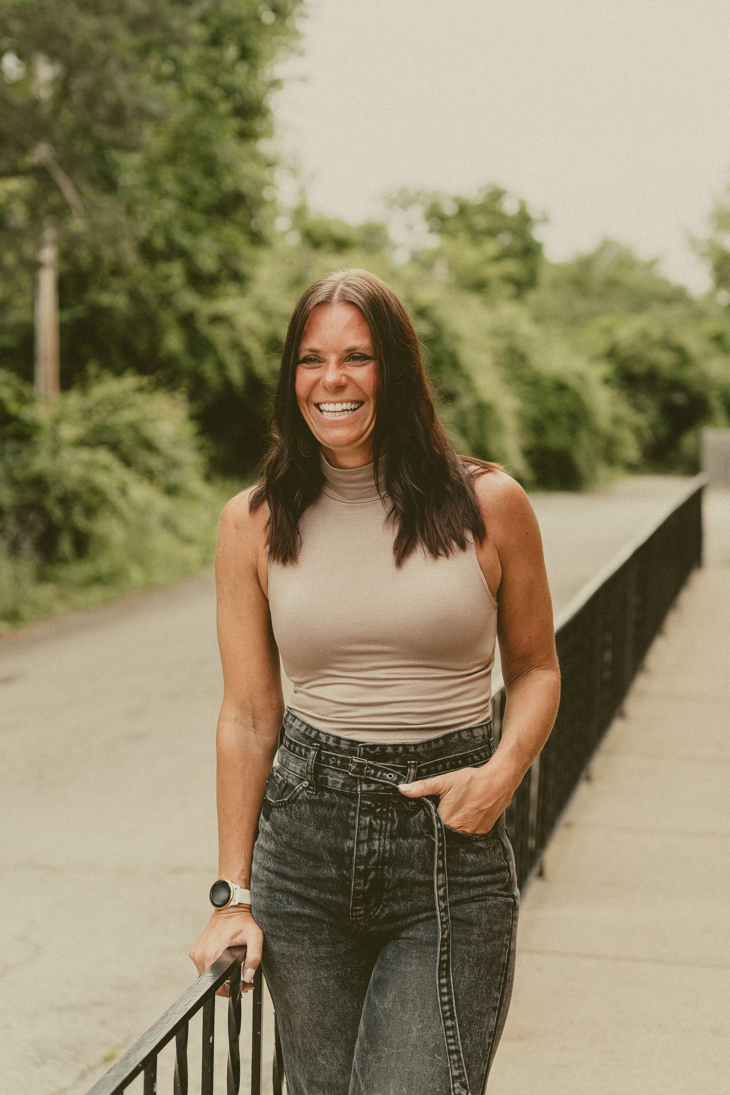 A person in a beige turtleneck and dark jeans poses casually along an outdoor railing in a natural setting at Brick and Beam Kettering Ohio.