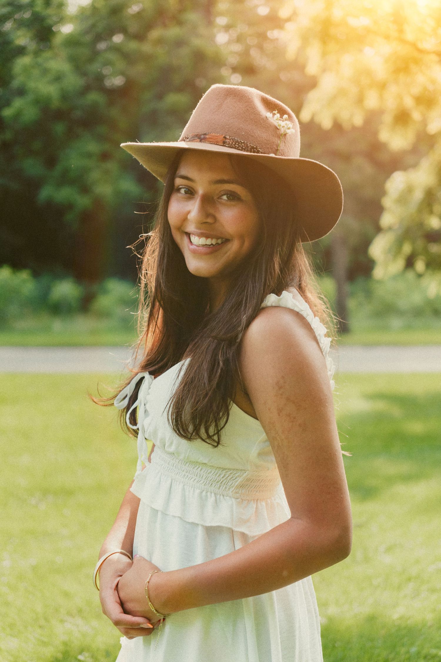 A girl in a white summer dress and brown hat poses playfully in a sunlit park setting in Bellbrook Ohio.