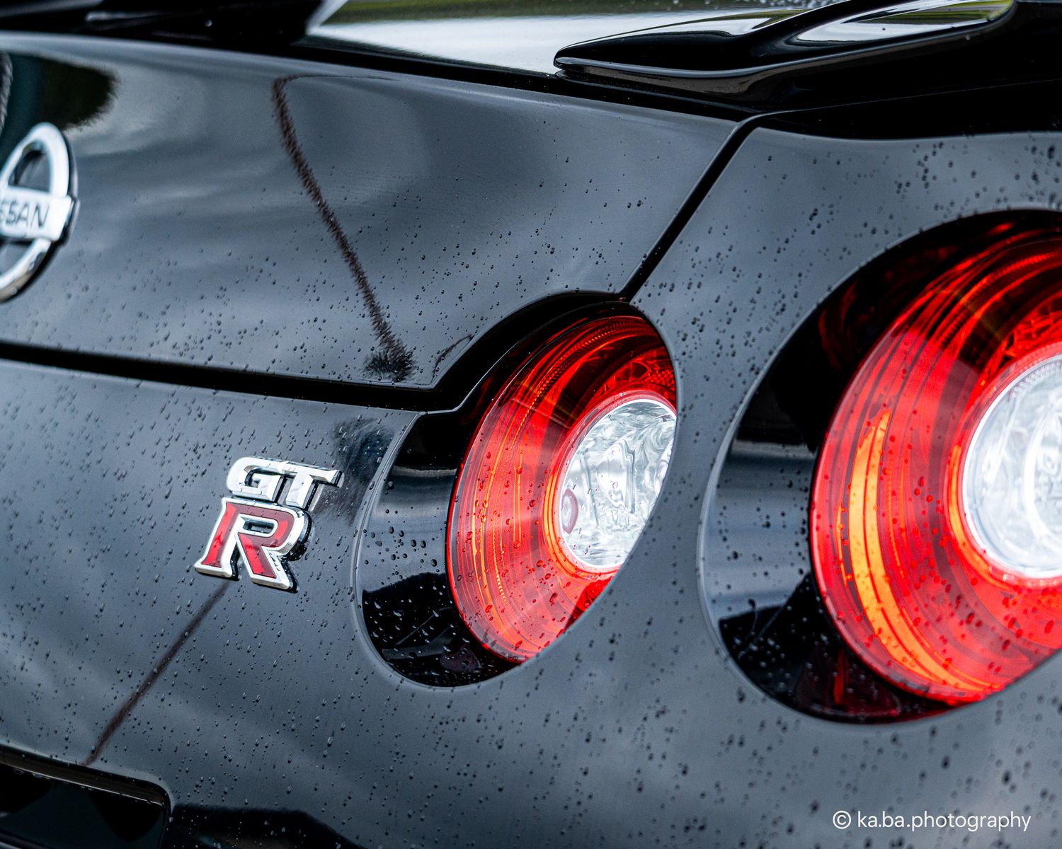 Close-up of Nissan GT-R rear badge and red tail lights with rain droplets on black paint.