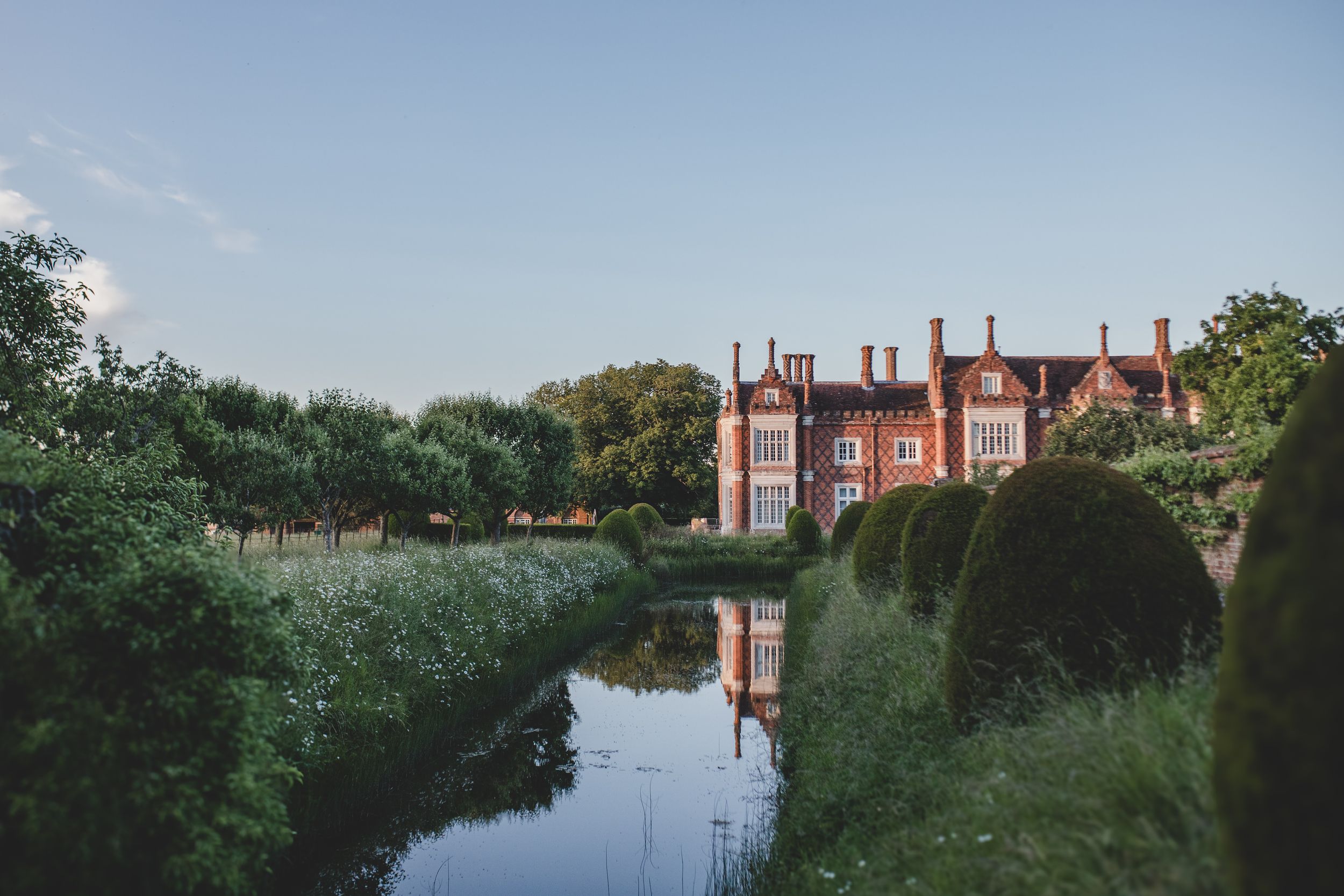 Historic red brick manor house reflected in a tranquil pond surrounded by manicured hedges and trees at sunset.