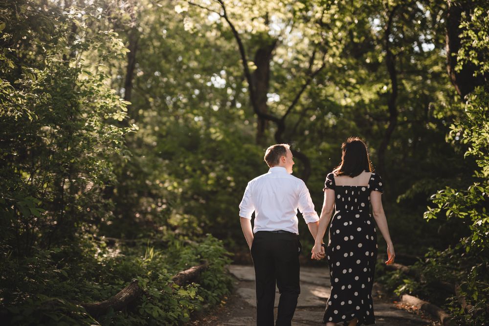 A couple walking hand in hand along a tree-lined path in a lush park setting.
