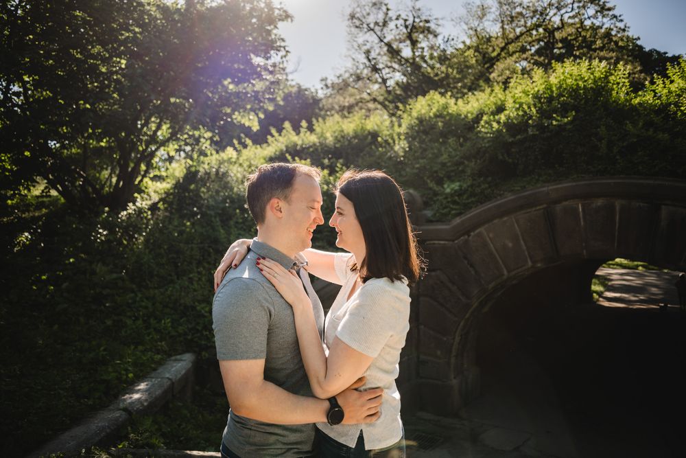 A couple embracing in front of a stone bridge with sunlight filtering through trees.