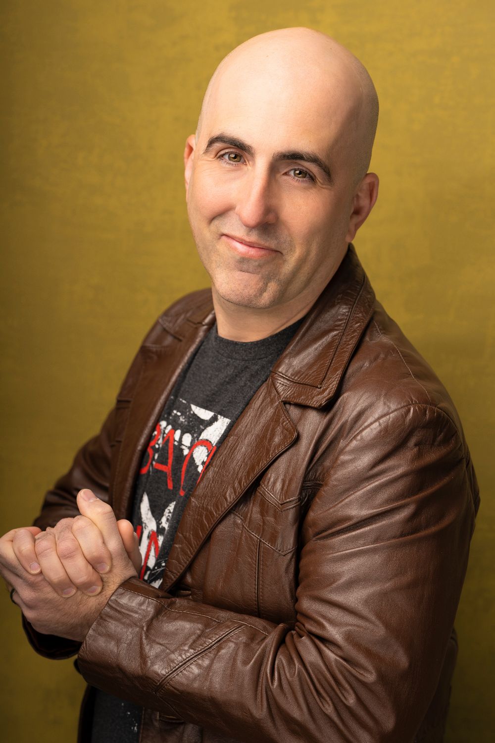 Professional studio portrait of person in brown leather jacket and graphic t-shirt against gold background.