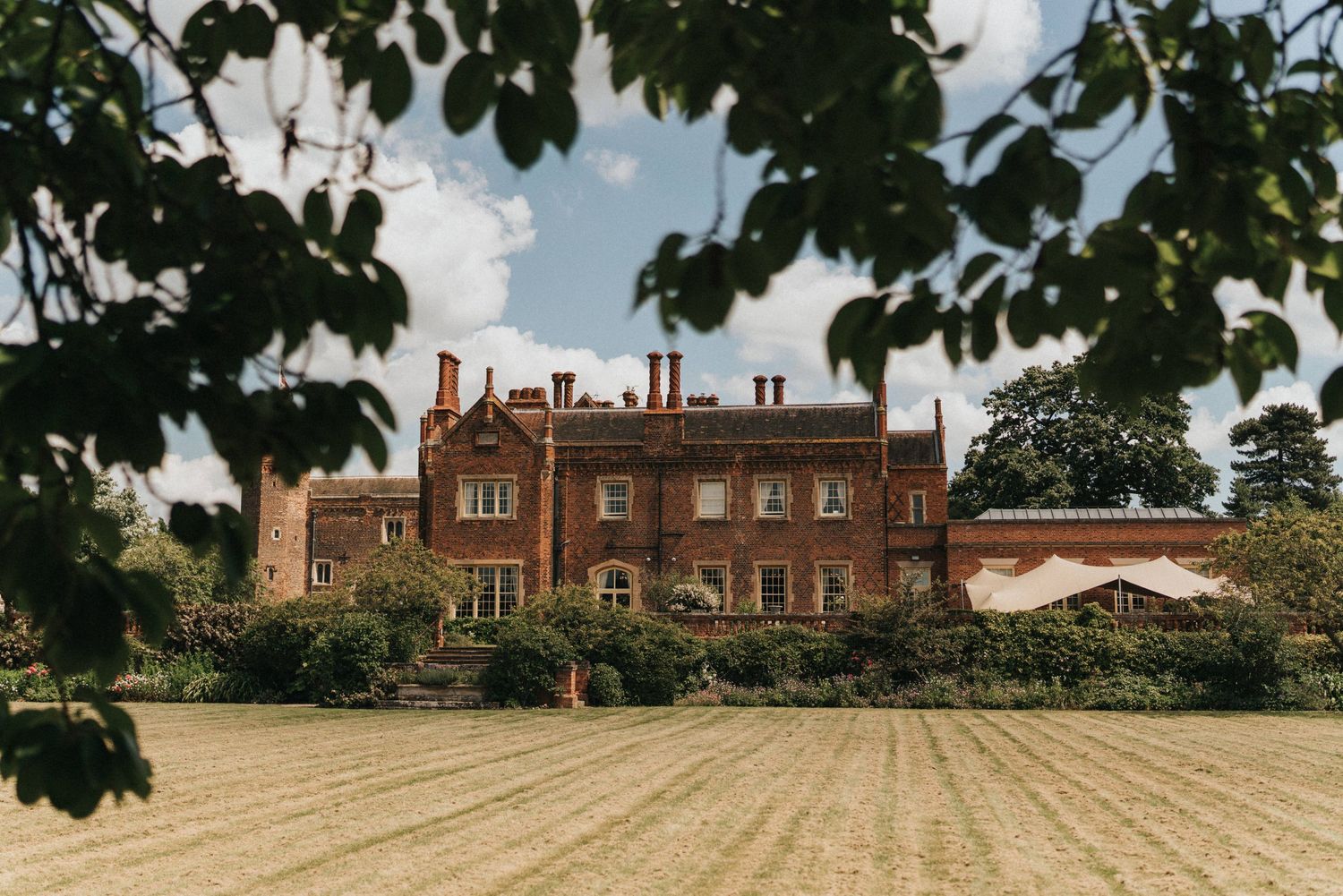 A panoramic view of a historic brick Tudor-style mansion with green landscaping and trees framing the architecture.