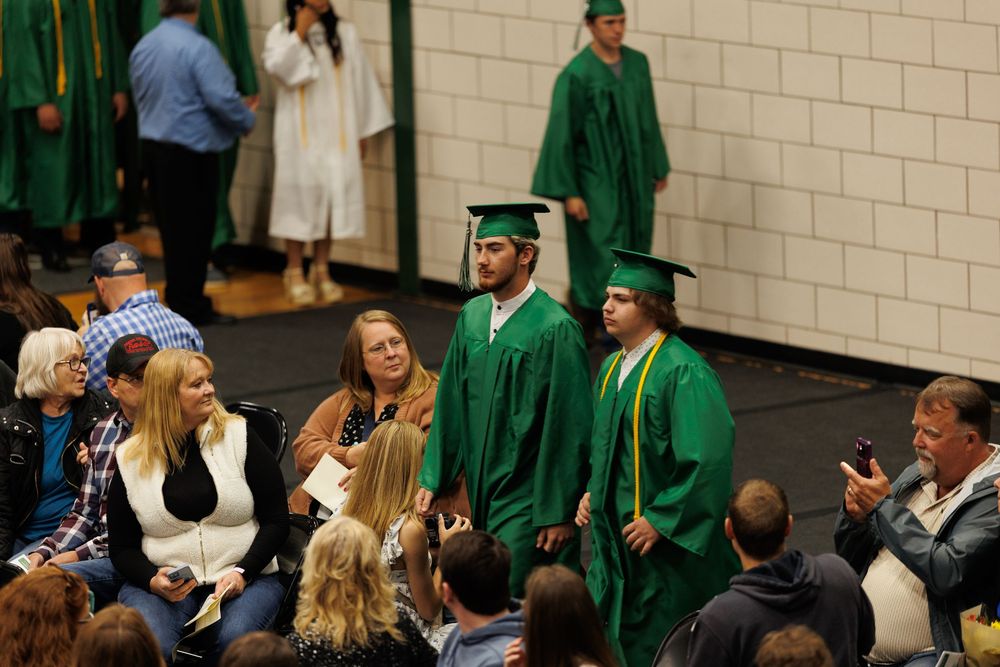 High school graduates in green caps and gowns receive diplomas at an indoor graduation ceremony.