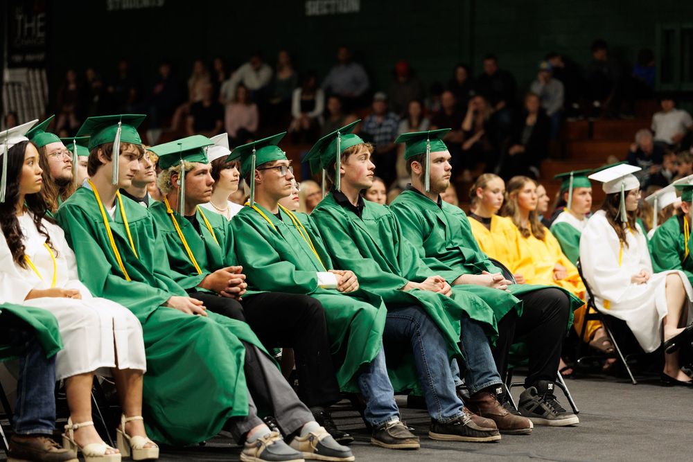 Row of graduates wearing forest green caps and gowns sit together during commencement ceremony indoors.