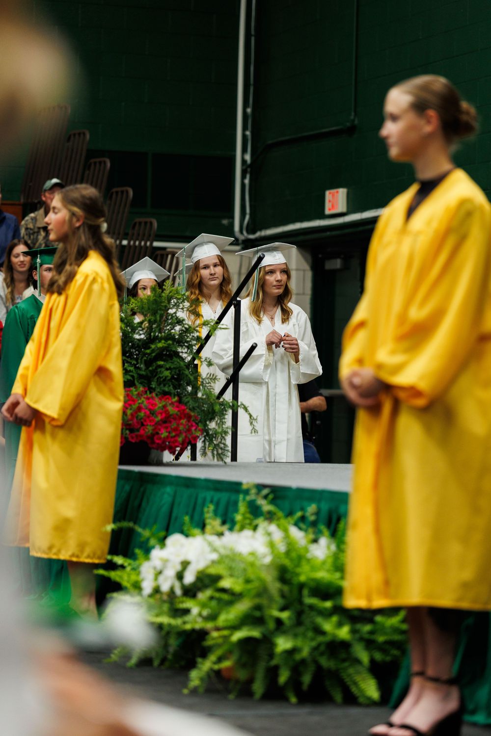 Graduates in bright yellow and white gowns stand on stage during a commencement ceremony with floral decorations.
