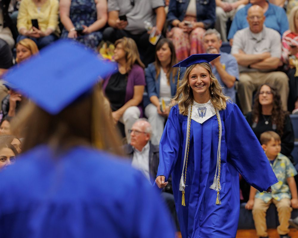 A student in bright blue graduation cap and gown smiles while walking during a commencement ceremony.