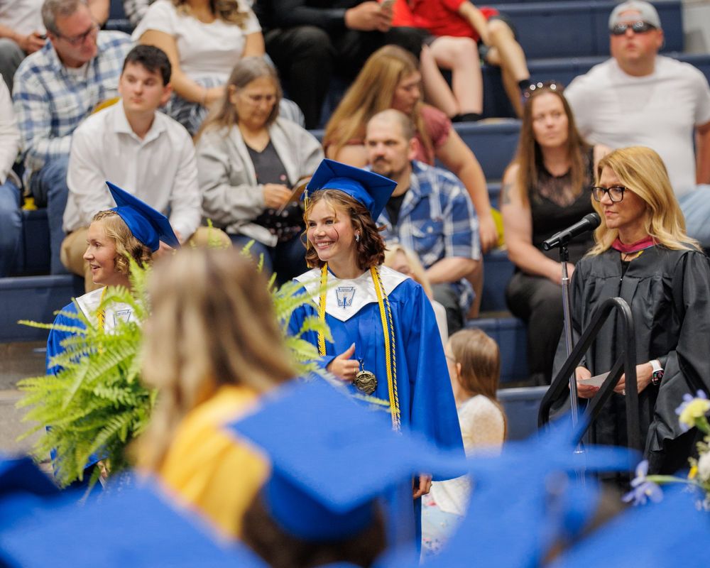 A row of graduates wearing blue and gold commencement robes sit during a graduation ceremony with audience in background.