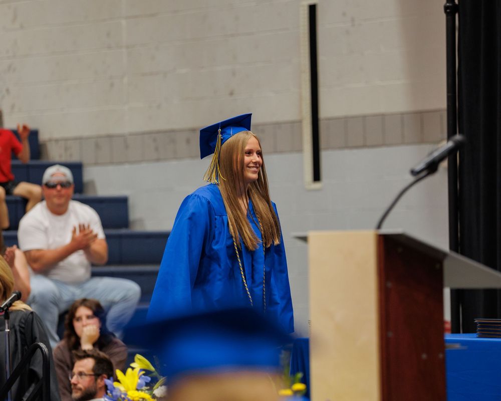 Graduate in bright blue cap and gown stands at podium during commencement ceremony while holding yellow flowers.