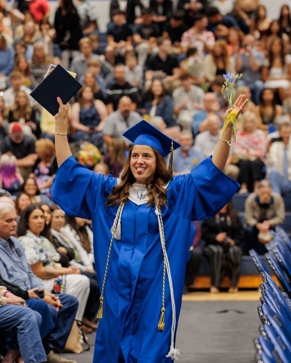 Graduate in blue cap and gown celebrating at commencement ceremony with raised arms holding diploma in crowded auditorium.