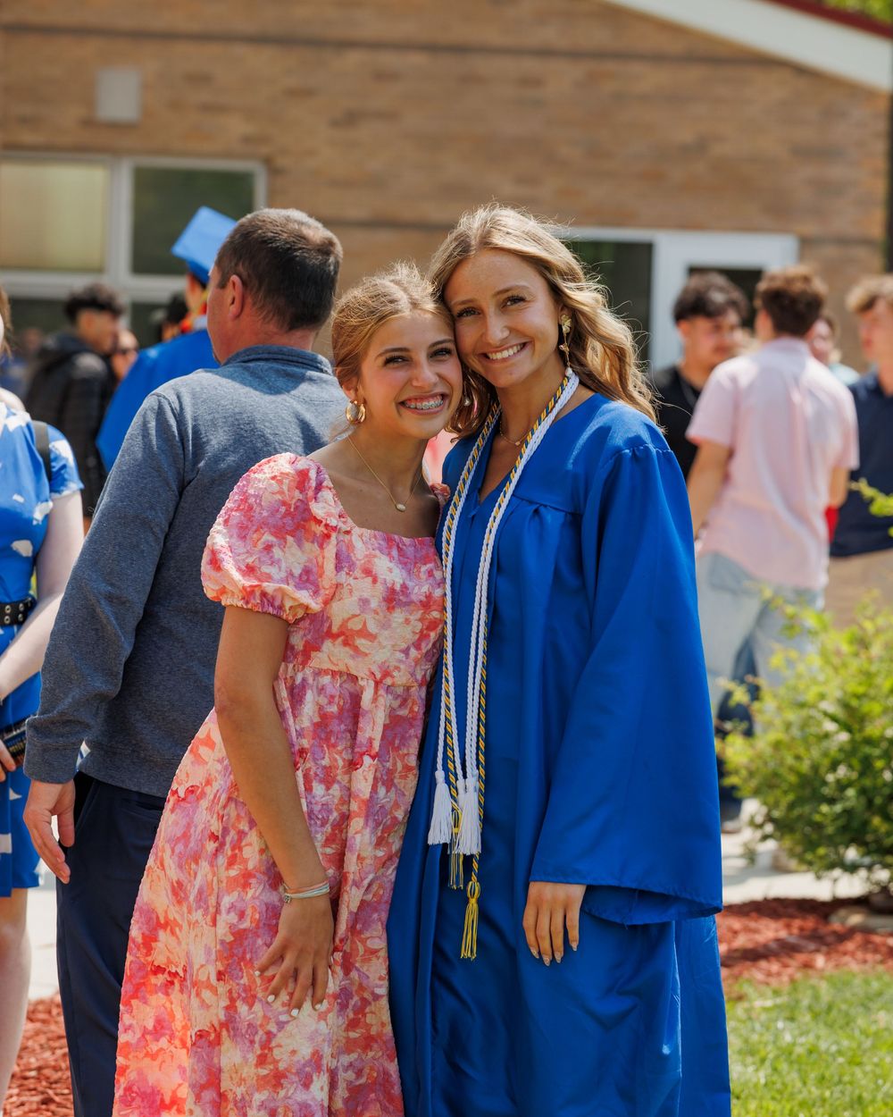 Graduates in blue robes and caps gather outdoors by brick building during commencement ceremony.