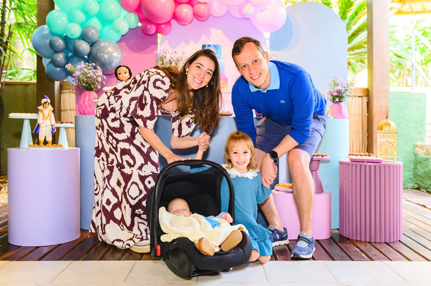 A family poses together at a colorful party setup with balloons in shades of pink, blue and mint green.