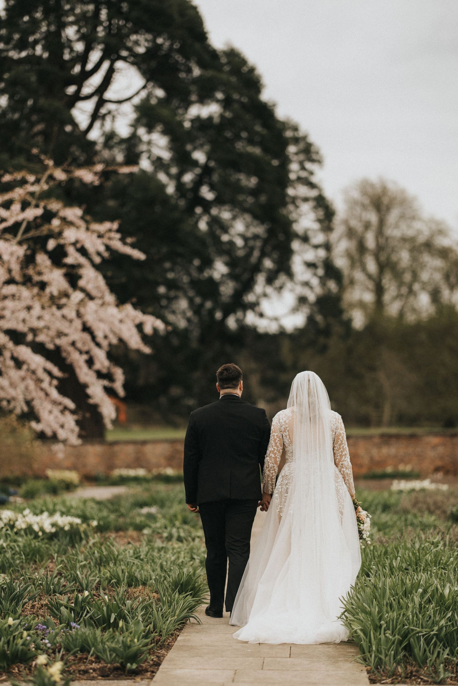 A wedding couple walks along a garden path surrounded by blooming cherry blossoms and spring flowers on a dreamy day.