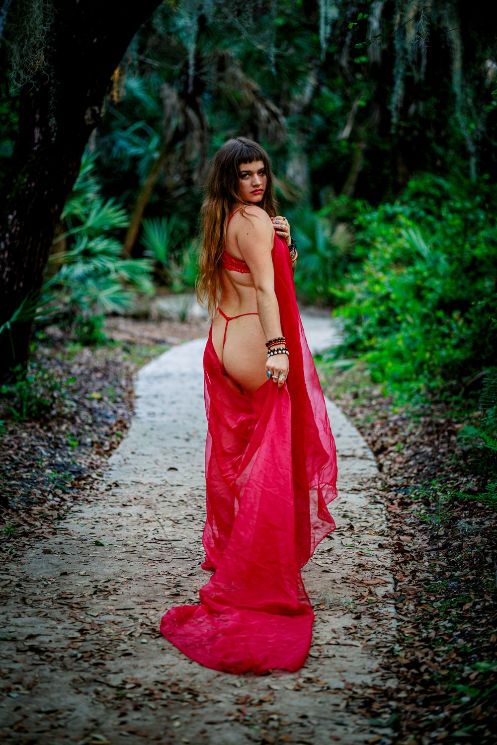 A figure in a flowing red dress poses gracefully on a forest path surrounded by lush green foliage.