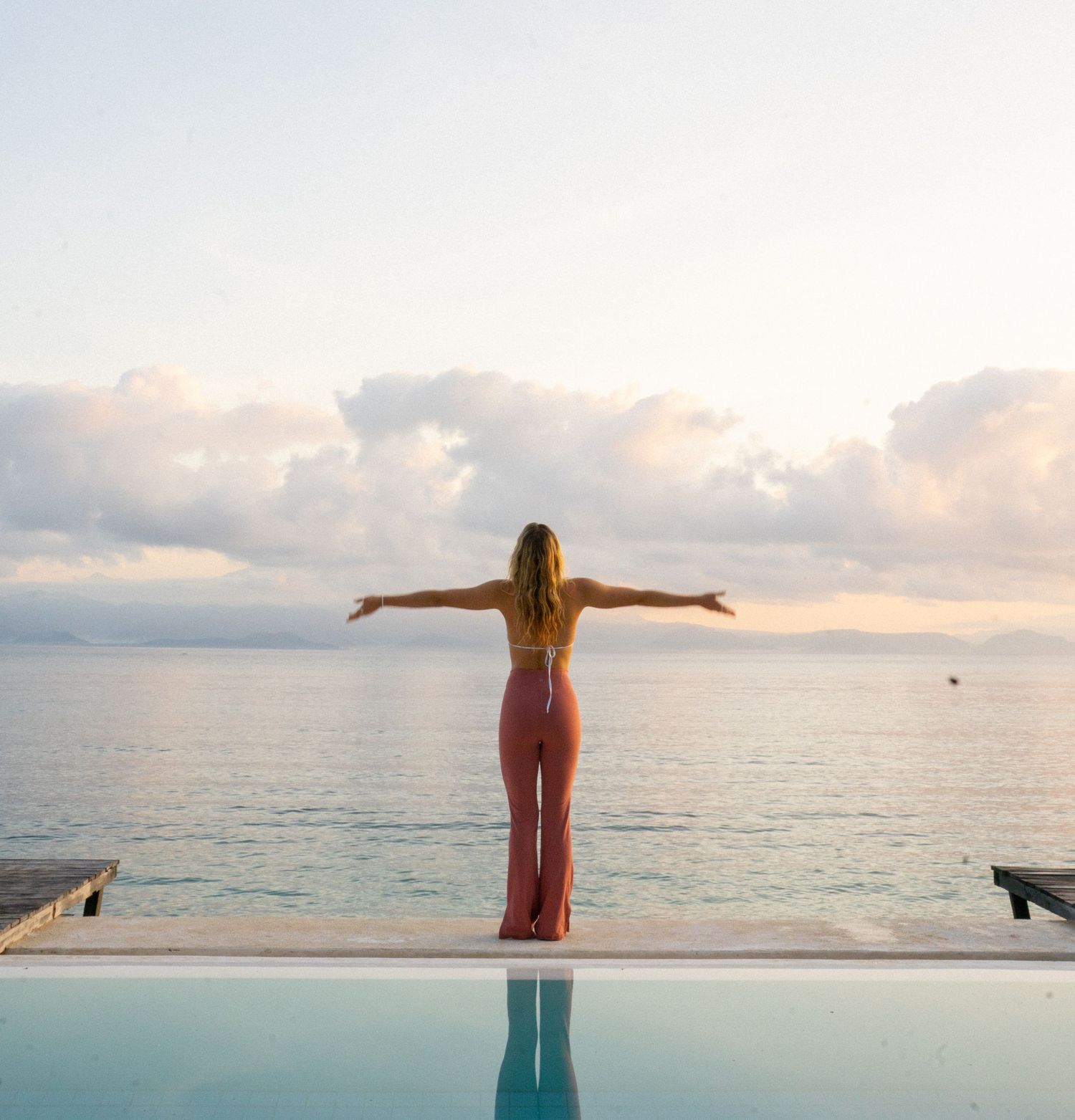 Silhouette of person standing with arms outstretched by infinity pool at sunset.