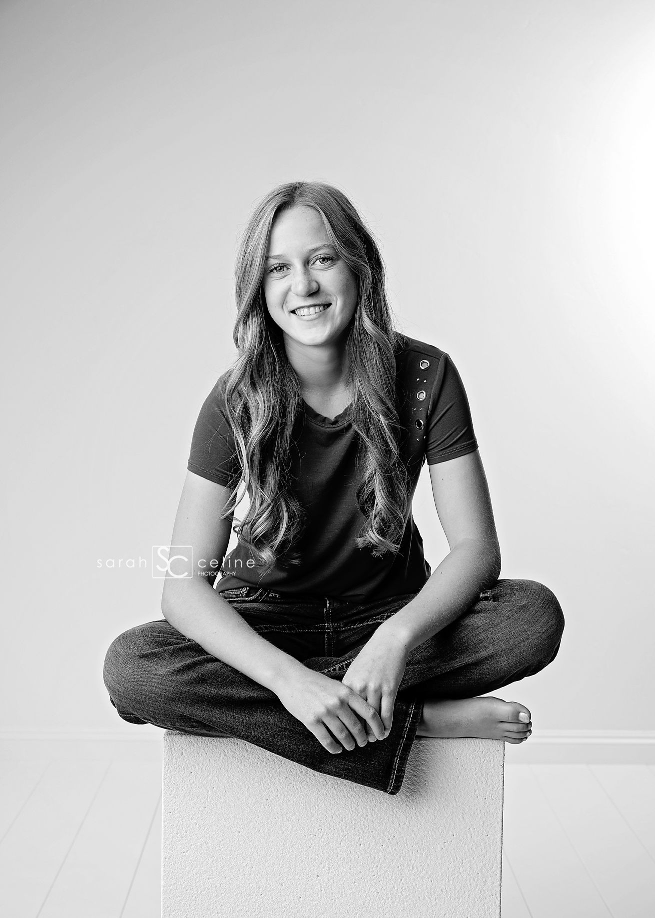 A black and white portrait shows someone in casual attire sitting cross-legged and smiling against a plain wall.
