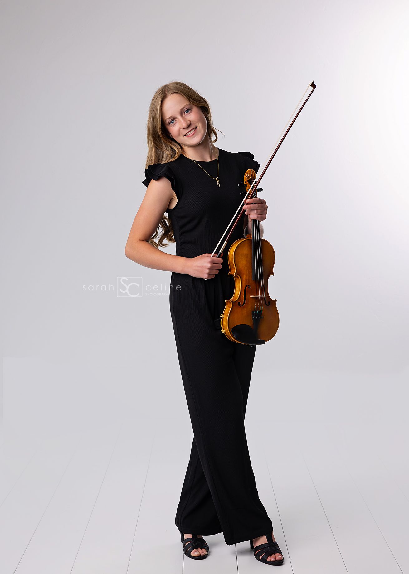 Young musician in elegant black attire poses with violin against studio backdrop.