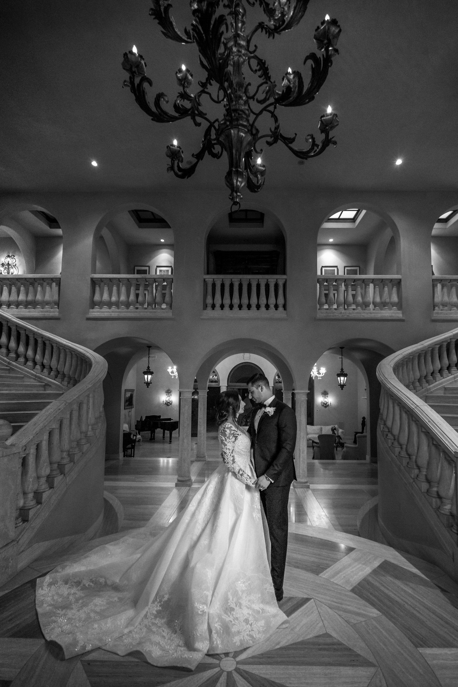 A romantic couple dances elegantly in a grand marble staircase with ornate chandeliers casting soft lighting in laguna beach estate.