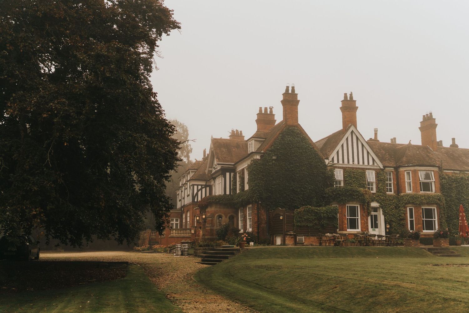 Healing Manor, a Vintage English Tudor-style mansion with brick facade and multiple chimneys stands on a misty lawn at dusk.