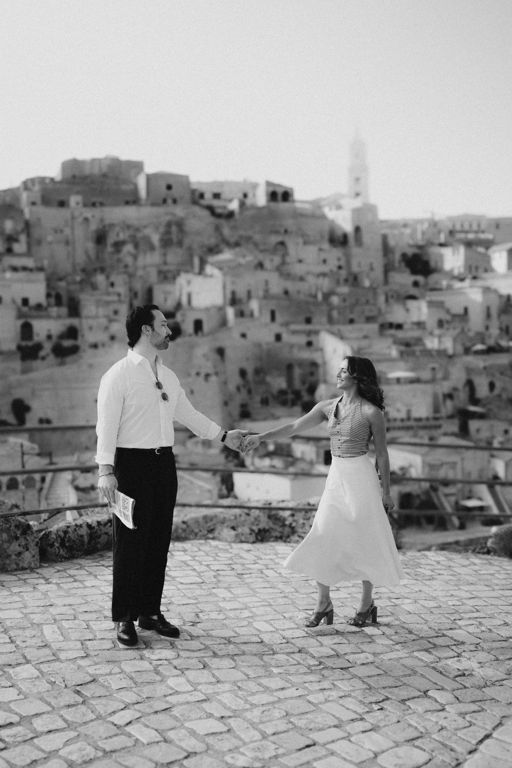 Two people stand on a cobblestone terrace against the backdrop of a historic hillside city in Italy.