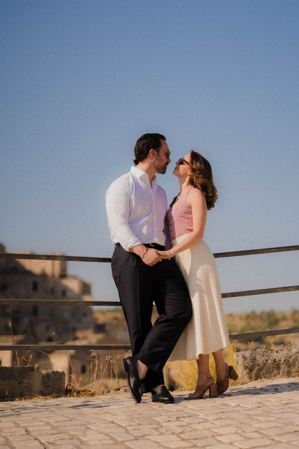 A couple in elegant attire share an intimate moment along a stone wall during golden hour.