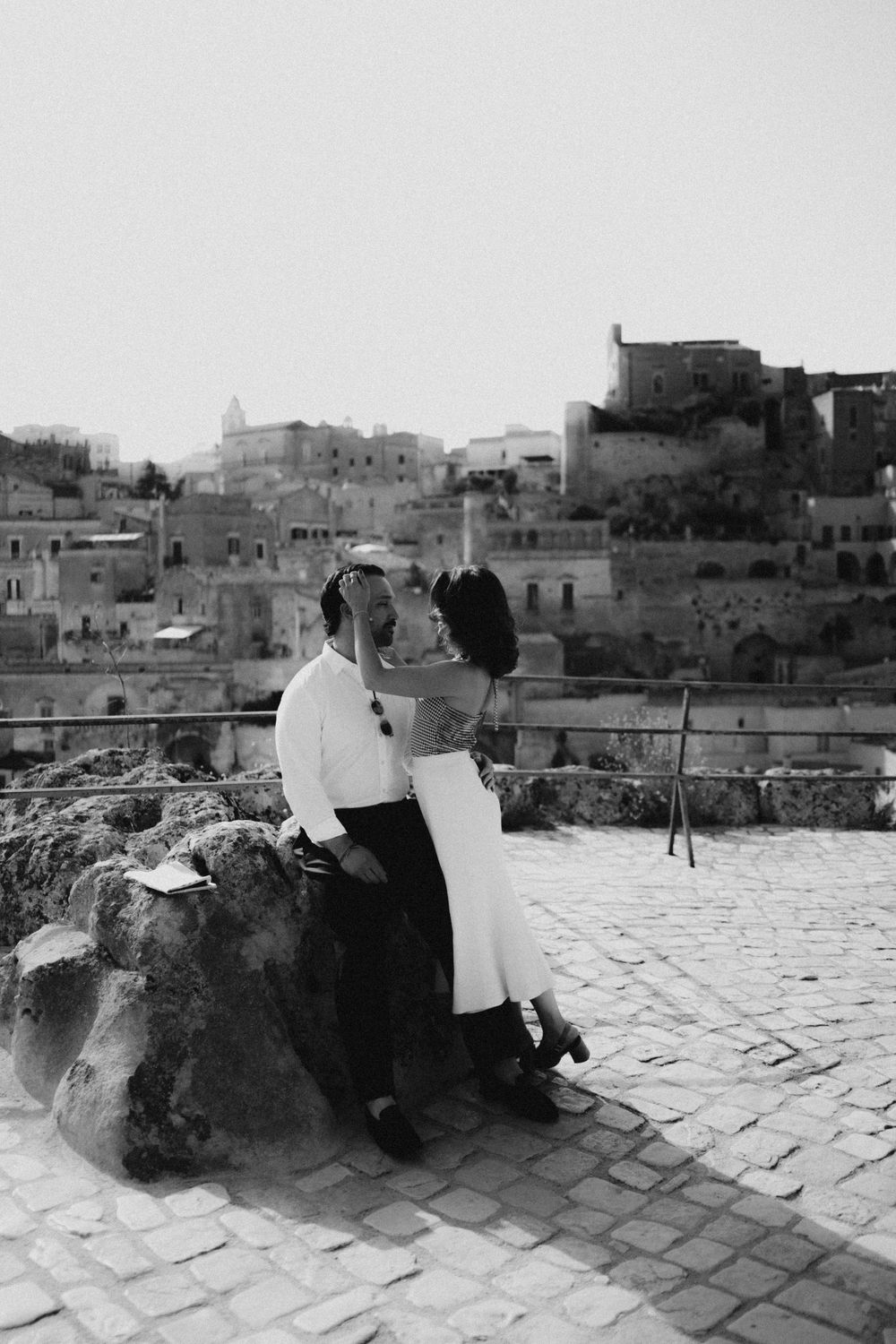 A romantic black and white photo of a couple sitting on stone steps overlooking an ancient Mediterranean town.