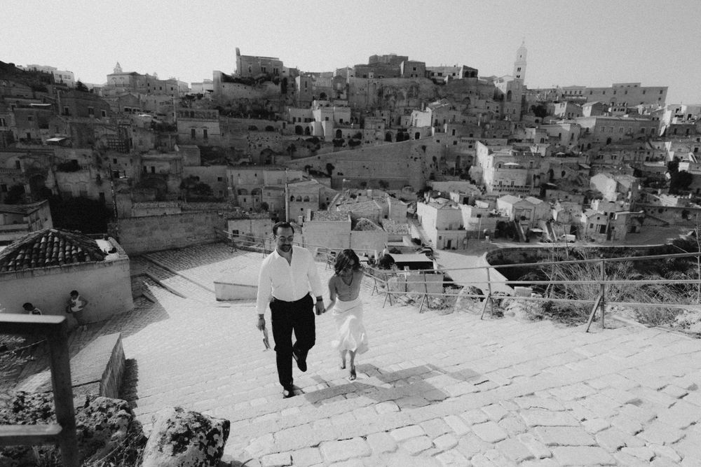 A black and white view of people walking down steep steps in a hillside town with clustered buildings in the background.
