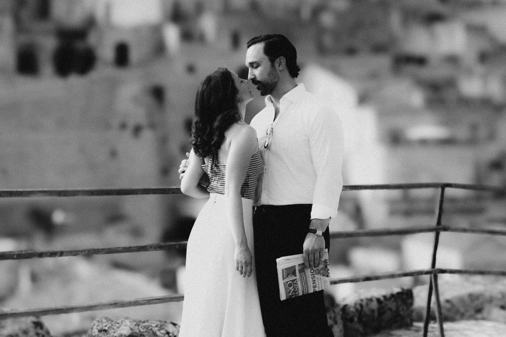A black and white portrait of a couple in elegant attire standing against a backdrop of historic buildings.
