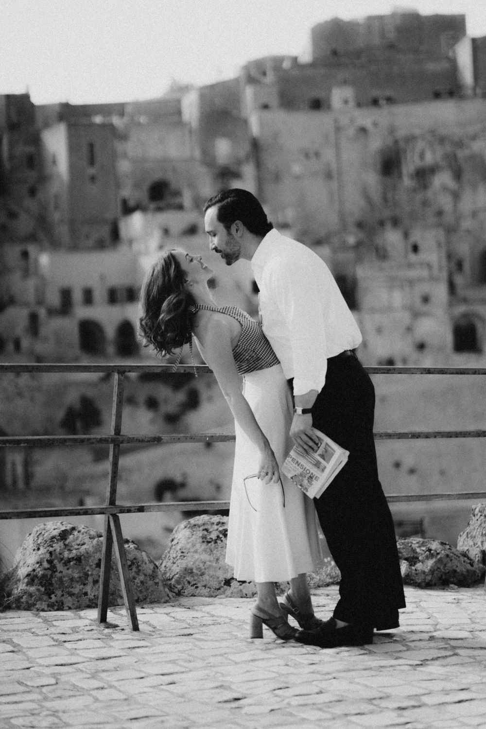 A couple in wedding attire shares an intimate moment on a terrace with a historic Italian village backdrop.