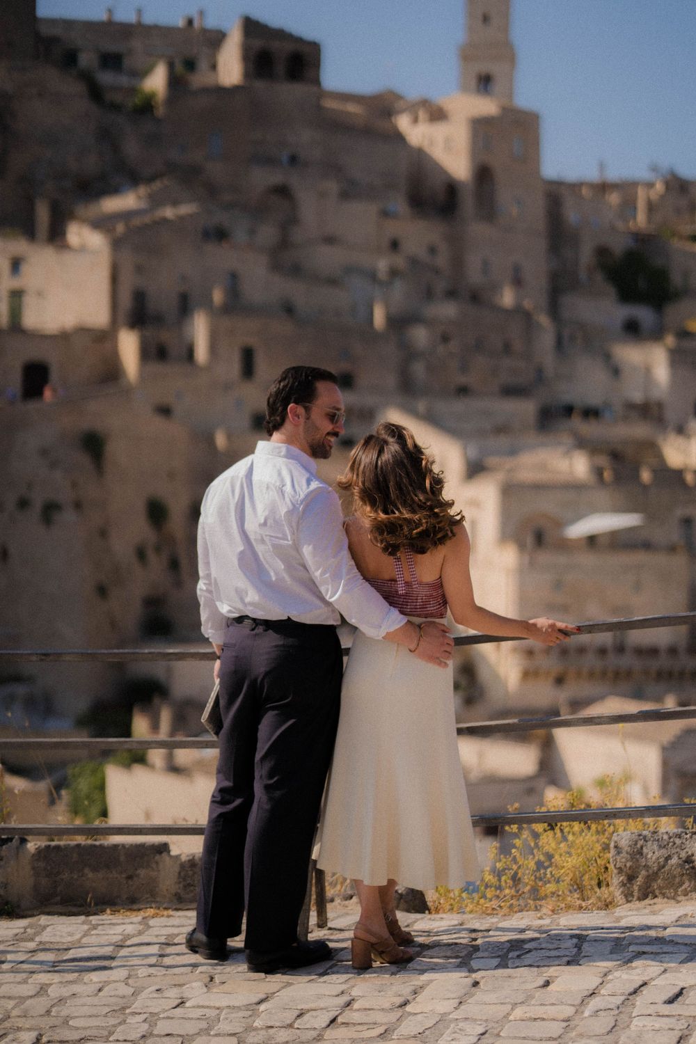 Couple sits on a stone wall overlooking the ancient cave dwellings of Matera, Italy on a sunny day.