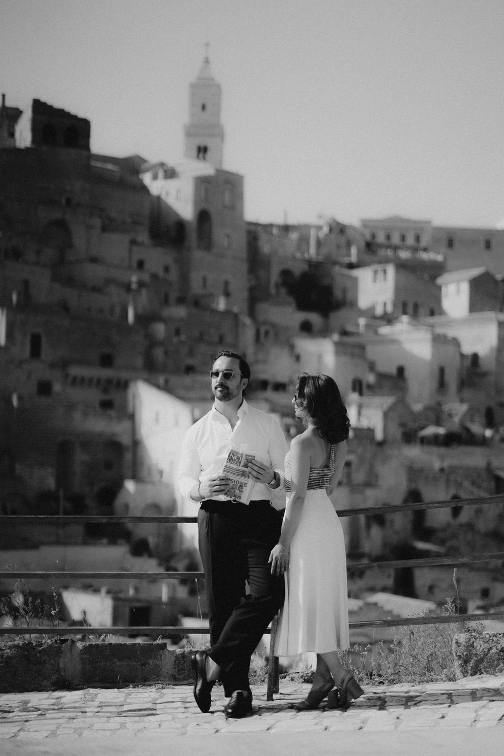 A romantic couple sits together on a terrace overlooking a Mediterranean town with white buildings and a church spire.
