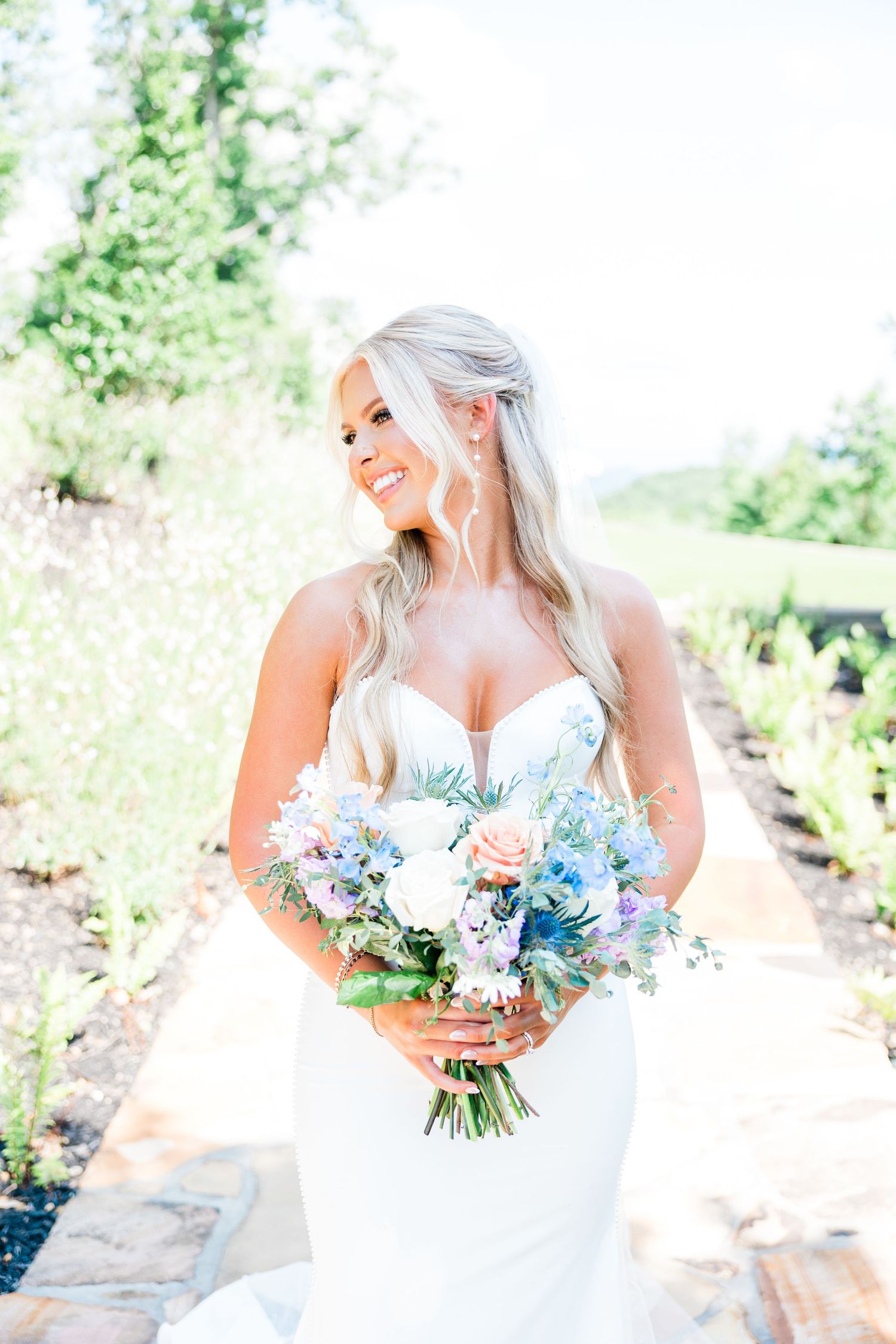 Ethereal bride in white gown holds pastel blue and white bouquet against natural outdoor backdrop.