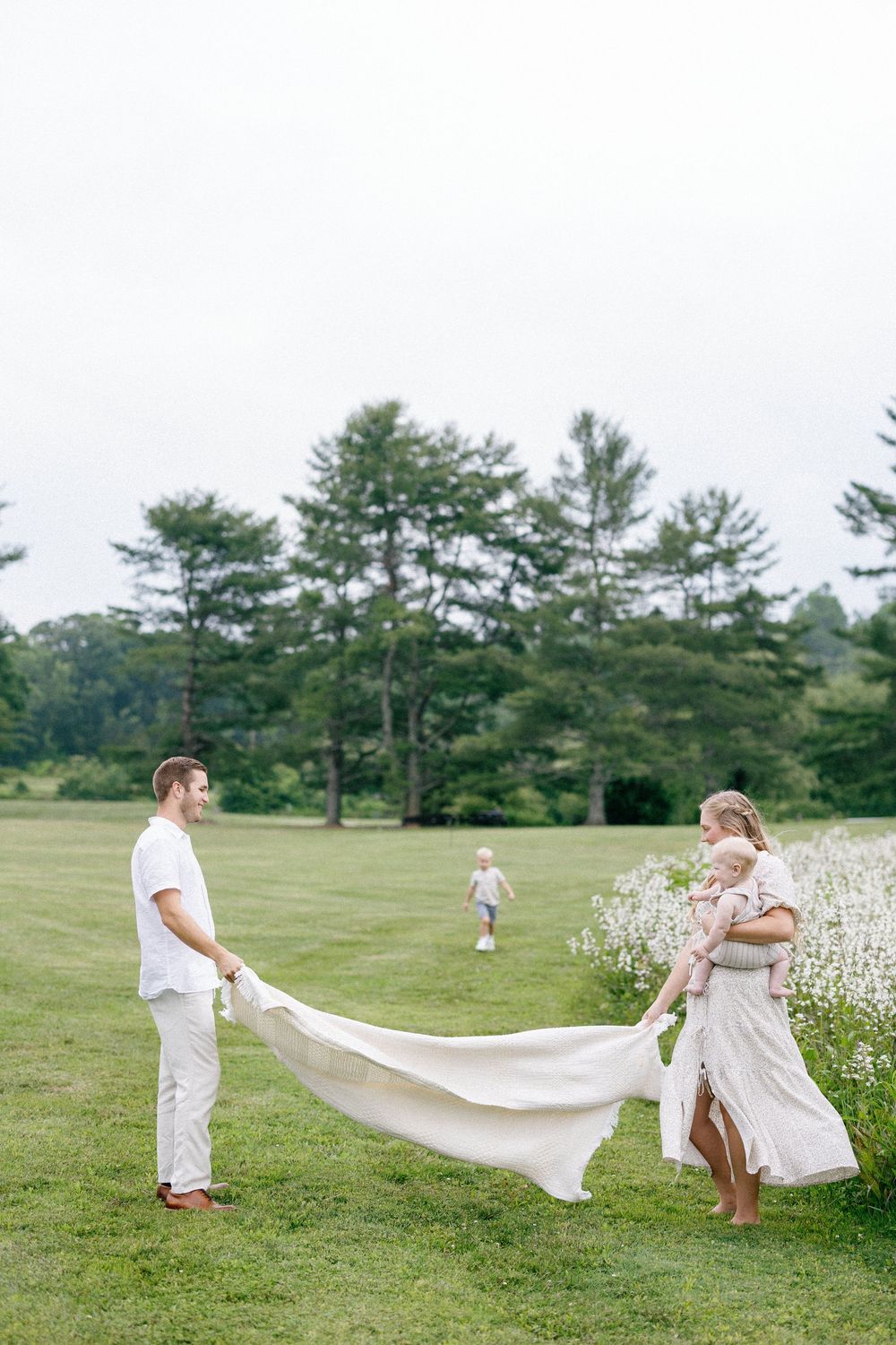 A couple holds the flowing train of a dress on a grassy field with pine trees in the background.
