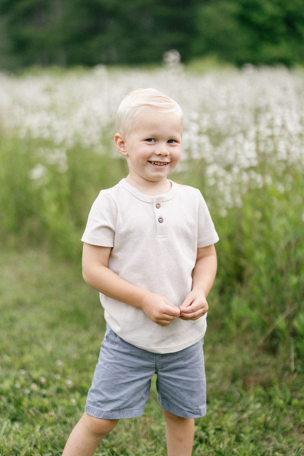 A child in a white shirt and grey shorts stands smiling in a field of white wildflowers on a sunny day.