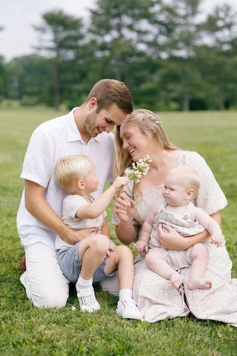 A tender family portrait on grassy field with toddlers and wildflowers in white summer outfits.