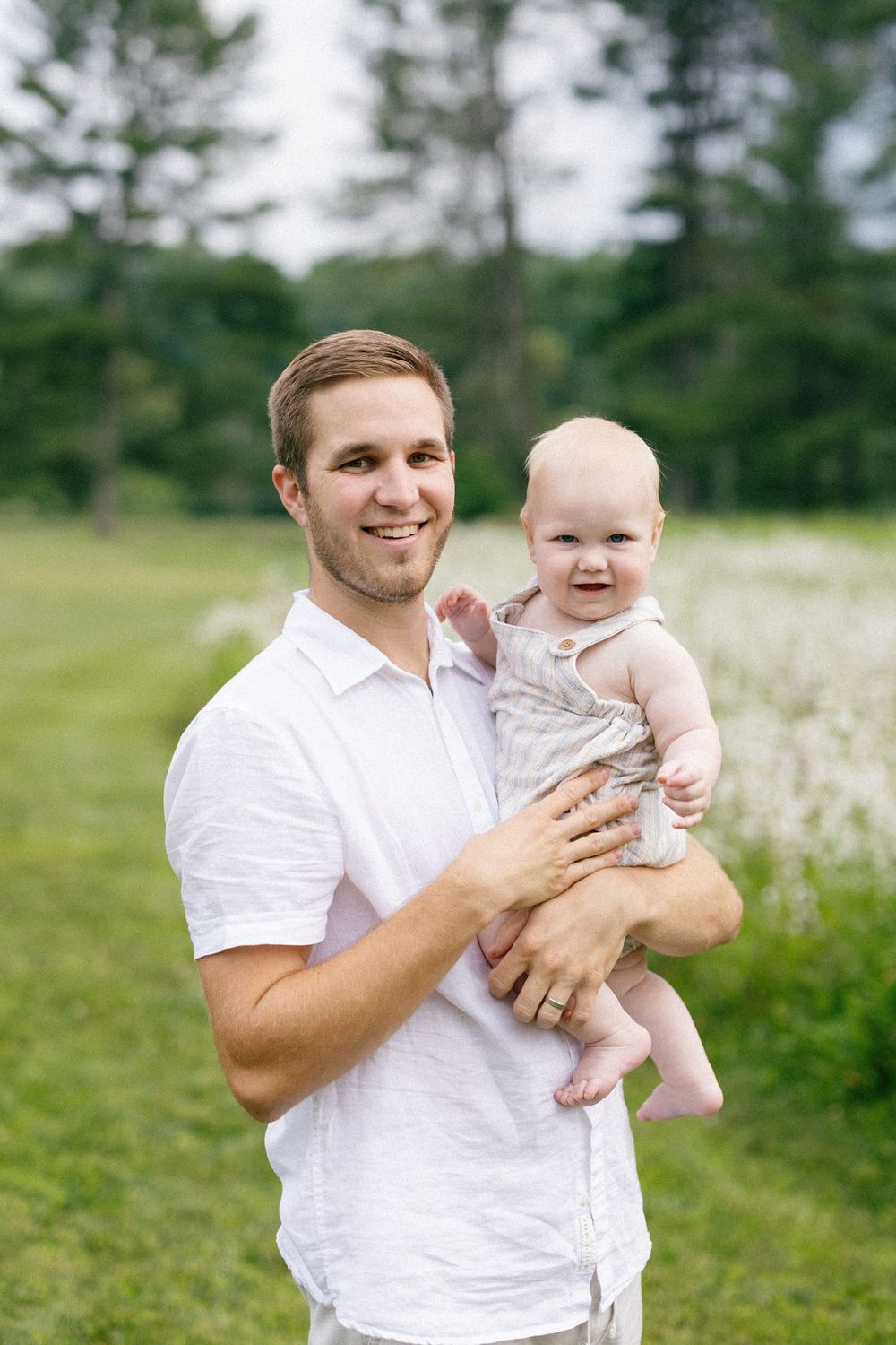 A joyful moment between parent and smiling baby against a backdrop of pine trees and meadow.