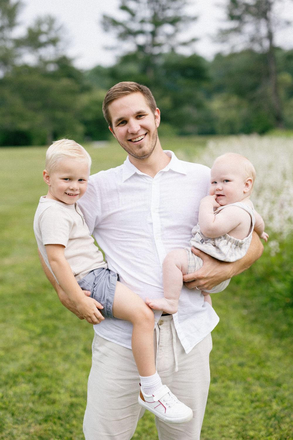 Outdoor family portrait in a grassy field showing a parent holding two young children on a sunny summer day.