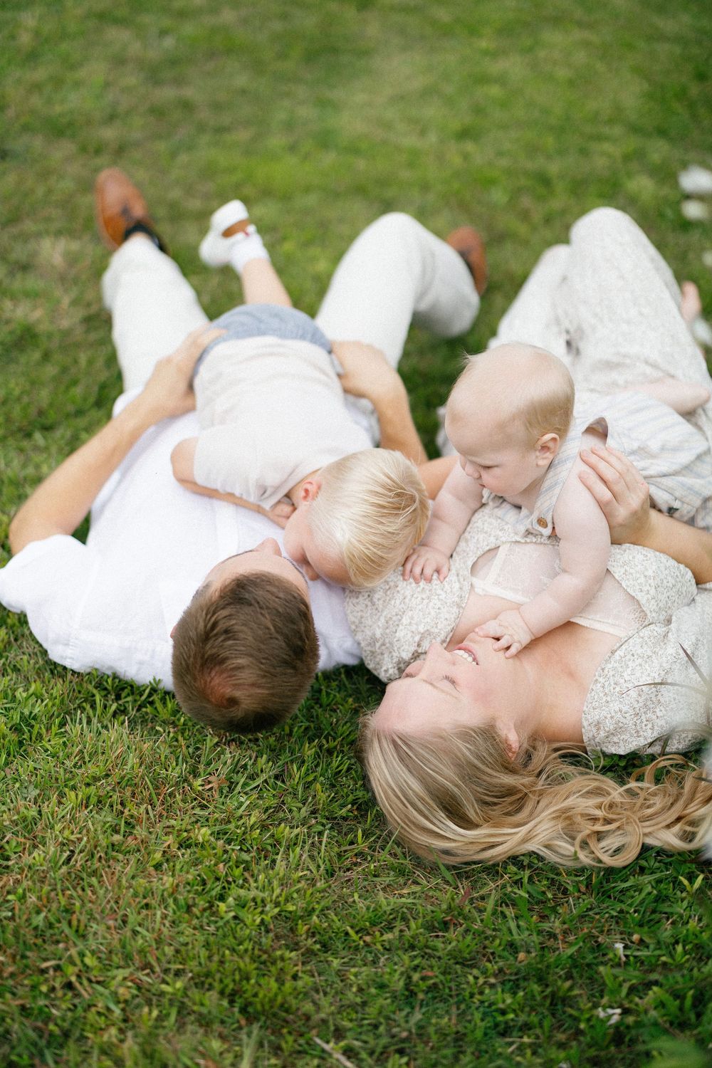 A family lying together on grass in coordinated white outfits during an outdoor photo session.