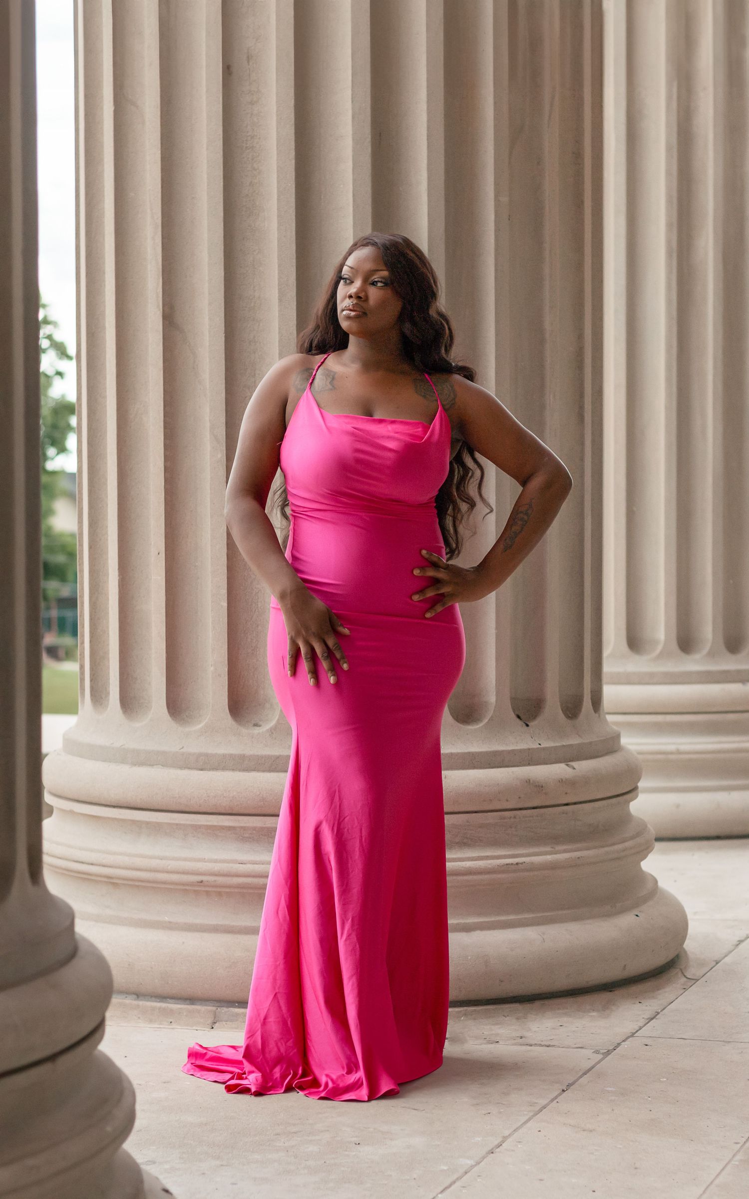 Model in a vibrant pink maxi dress poses elegantly among classical stone columns in an architectural setting.