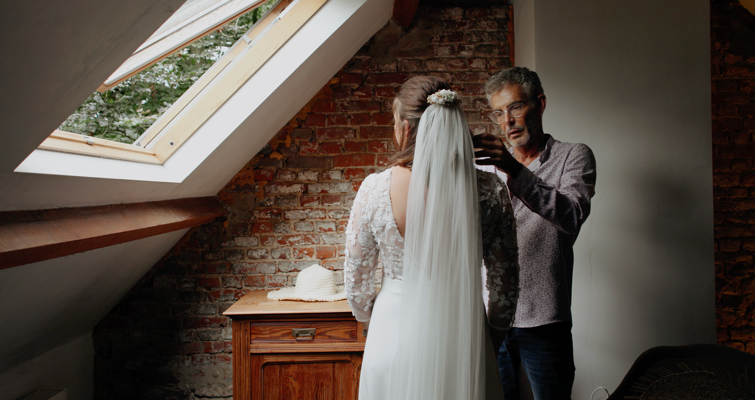 A person adjusts a wedding dress on display in a cozy attic room with exposed brick walls and skylight window.
